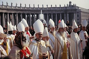 Para uskup berbaju putih berdiri di bawah sinar matahari di Lapangan Santo Petrus. Sebagian besar mengenakan mitra putih di kepala mereka, kecuali seorang uskup Katolik Timur yang mengenakan topi beludru bersulam yang khas.