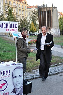 Libor Michálek in a suit walking near another man and his 2012 Senate campaign booth, which has a white background with purple and magenta lettering: "Michálek do Senátu" and a whistleblower logo (a metal pea whistle with a slanted exclamation point above, surrounded by a circle)