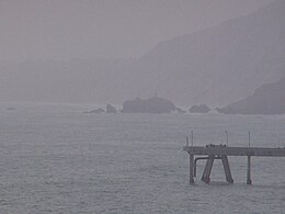 Ein einsamer Pier ragt vom rechten Rand in ein Foto hinein; Im Hintergrund sieht man durch einen melancholischen Nebel mehrere unscheinbare, gezackte Felsen, die sich aus dunkelgrauem Wasser erheben.