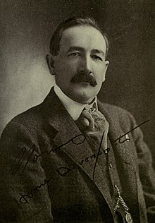 Photographie en noir et blanc représentant le portrait d'un homme arborant une fine moustache.