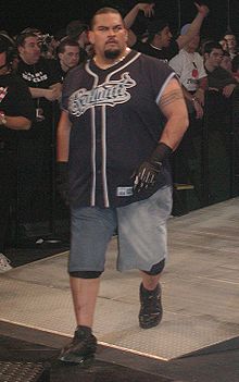 Un luchador en la rampa avanza hacia el ring.  Lleva una camiseta similar a la de un equipo de béisbol, pantalones capri de mezclilla, zapatillas negras, guantes negros y un collar de oro.  Tiene el pelo rapado.