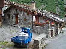 De kleurenfoto toont een kleine blauwe gemotoriseerde scooter beladen met 4 aluminium melkkannen.  Het podium bevindt zich in een dorp met stenen huizen en stenen platen en leien daken.