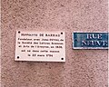 Plaque de couleur blanche à écriture noire rendant hommage à Hippolyte de Barrau pour la fondation de la Société des lettres, sciences et arts de l'Aveyron. À sa droite, une plaque bleue indiquant la « rue Neuve ».