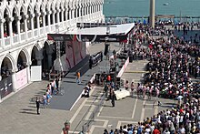 A large crowd of spectators gathers behind barricades to observe a cycling team be publicly presented on an elevated dais