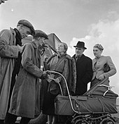 two white men in flat caps, with a pram, talking to middle aged couple and young woman