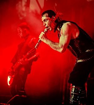 Lindemann ist hinter einem rot gefärbten Bühnenbild abgebildet. Er hält ein Mikrofon und trägt dunkle Kleidung.