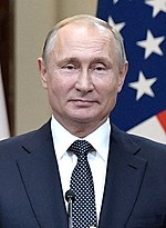 in front of American flag, chest height portrait of smiling man in suit and tie with dyed thinning hair in his sixties