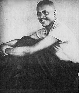 Photographie en noir et blanc d'un homme assis sur le sol. Ses jambes sont pliées et ses bras entourent ses genoux. Sa tête est tournée vers sa gauche et il sourit.