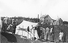 Deux femmes et quatre enfants prennent la pose devant une tente de toile. Derrière la tente, une corde à linge sur laquelle pendent des vêtements, et une cabane de bois.