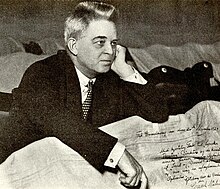 Foto von Carl Nielsen in dunklem Anzug und Krawatte, der hinter zerknitterten Papieren sitzt, darunter ein Brief mit seiner Unterschrift