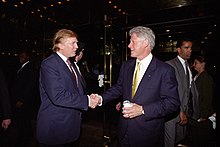 Donald Trump aperta a mão de Bill Clinton em um saguão;  Trump está falando e Clinton está sorrindo, e ambos estão vestindo ternos.