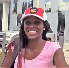 Headshot hitam wanita mengenakan topi baseball dengan Mozambik bendera di atasnya, dengan kaca gedung dan mobil putih di belakangnya