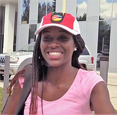 Headshot of a black woman wearing a baseball cap with a Mozambique flag on it, with a glass building and white car behind her