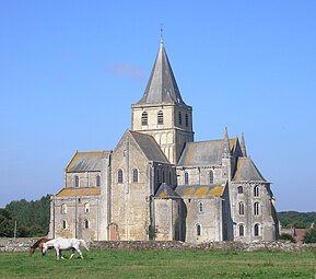 Uma alta igreja de pedra cinzenta com detalhes finos e uma torre de travessia encimada por um pináculo coberto de ardósia ergue-se do campo rural, onde duas éguas pastam.