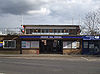 Um edifício de tijolos marrons com uma placa azul dizendo "GRANGE HILL STATION" em letras brancas, tudo sob um céu azul com nuvens brancas