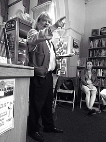 foto di un uomo allegro e barbuto che indica il pubblico con una mano mentre legge dal libro delle sue poesie con l'altra.