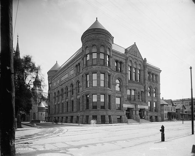 A black-and-white photo of a four-story stone building from across an empty street; a church stands off to its left and several two-story houses sit to the right down the street.