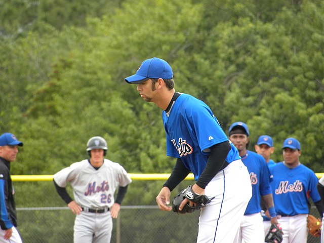 A man in a blue baseball jersey and baseball cap and white baseball pants bending over from the waist; he wears a black baseball glove on his left hand