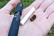On an open palm, a glossy plastic IQOS holder, the short paper-wrapped mini-cigarette that was in it, and the somewhat darkened tobacco plug removed from the end of the paper cylinder.