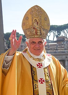 Frontale Farbfotografie vom Papst mit einer gelben liturgischen Kleidung und einer rot-weißer Stola. Die Kopfbedeckung zeigt eine Szene mit einem Heiligen und einem Kreuzstab, der von zwei Lämmern umgeben ist.