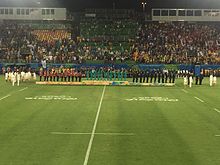 vista da arquibancada em frente ao pódio olímpico, com os jogadores todos voltados para a direita.