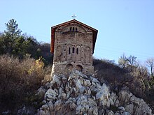 Ostansicht einer schmalen mittelalterlichen Kirche auf einer Klippe mit einigen Wohngebäuden im Vordergrund. Die Apsis der Kirche ist hinter einigen Felsen sichtbar.