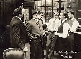 In a still from The Corner (1916), millionaire David Waltham (played by George Fawcett, holding a cigar and scowling) stands in a group of businessmen.