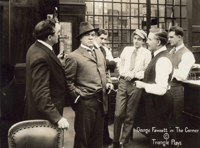 In a still from The Corner (1916), millionaire David Waltham (played by George Fawcett, holding a cigar and scowling) stands in a group of businessmen