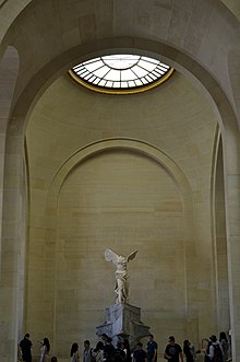 Scultura in marmo raffigurante una donna alata in piedi vestita di toga, senza braccia né testa.