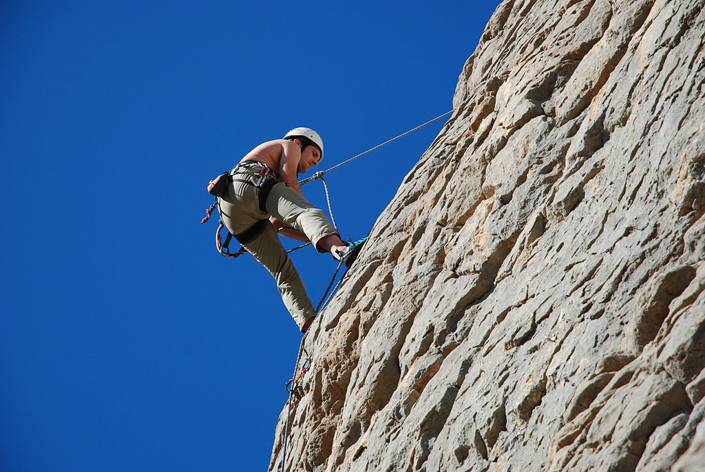 Topless man abseiling down a scraggly rock face with a rope. Blue sky in the background.