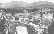 Birger Ruud beim Sprung, mit dem Zielgelände und den Bergen im Hintergrund