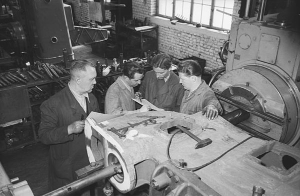 Photograph of East German machine tool builders in 1953, from the German Federal Archives. The workers are discussing standards specifying how each ta