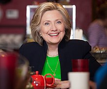 Хиллари Клинтон в черном костюме и зеленой рубашке сидит в кафе. Она улыбается, и перед ней стоит красная чашка. Передний план искажен из-за наличия различных мелких предметов. 