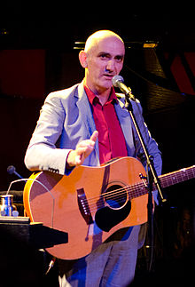 56-year-old Kelly is standing at a microphone with his guitar slung over his shoulders. His right arm is bent at the elbow towards the viewer, while his left is at his hip. He wears a grey suit with an orange shirt.