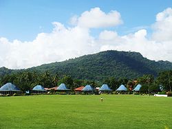 Lepea village with round meeting houses where matai meetings take place and open commons area (malae) for outdoor ceremonies. Round Samoan houses with blue roofs, Lepea village with Mt Vaea beyond.JPG