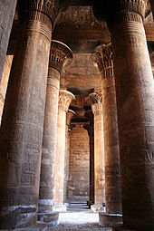 Large, shadowy room filled with tall, thick columns. The column capitals are shaped like stylized flowers.