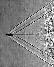Shadowgram of shock waves from a supersonic bullet fired from a rifle. The shadowgraph optical technique reveals that the bullet is moving at a Mach number of about 1.9. Left- and right-running bow waves and tail waves stream back from the bullet, and its turbulent wake is also visible. To the right of the frame are seen unburned powder particles from the cartridge, themselves traveling slightly subsonic so that each of them produces a curved bow shock wave.