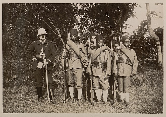 un officier en uniforme bleu foncé au côté de six tirailleurs malgaches en uniforme kaki