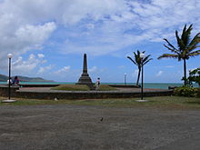 A stretch of a bay. In the foreground is a gravel area in front of a large obelisk and two palm trees.