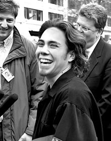 A black and white photo featuring a joyous man with a large smile who is giving an interview to a group of smiling news reporters that are crowded around him.