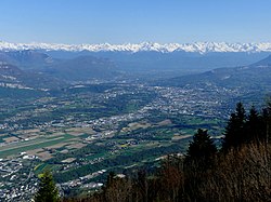 Vue de la cluse de Chambéry depuis le mont du Chat au nord avec Chambéry et son agglomération au centre, le massif des Bauges à gauche, le massif de la Chartreuse à droite et la chaîne de Belledonne enneigée par delà la combe de Savoie au loin.