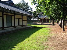 Em um dia ensolarado, um edifício de madeira tradicional coreano pintado de branco e vermelho escuro fica em um campo de grama.  Árvores luxuriantes são vistas à direita, enquanto um portão é mostrado à distância.