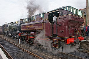 A pannier tank locomotive is standing at a platform, leading a train which includes a second locomotive and one visible passenger carriage. The pannier tanker is maroon, apart from the black chimney and red coupling rods and buffer beam. The tank, cab, steps, splashers and toolbox are all lined in yellow. London Transport is written on the side of the tank, and L.99 on the side of the cab, again in yellow. The second, larger, locomotive is green with a brass safety valve cover, a name plate over one of the wheels, and has a tender behind it.