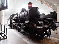 Sort dampeksprestogslokomotiv som holder i en stor lys remise. Lokomotivet er poleret op, og er iført rødt og hvidt skorstensbånd.