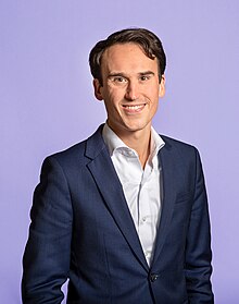 A smiling Caucasian man with dark hair in a suit in front of a purple background