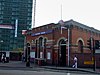 Un edificio en forma de cubo de ladrillos rojos con un letrero rectangular de color azul oscuro que dice "PLAISTOW STATION" en letras blancas y gente caminando en la acera