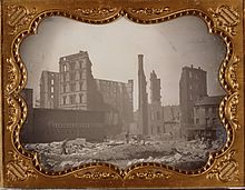 Ruins of the Lindell Hotel after the fire of 30 March 1867. Men work amongst the rubble and a horse can be seen near a building off to the left. Portions of the building still stand in the background. A brick structure next door is labeled with a sign that reads "Lindell Livery Stables", and a man leans up against it. Ruins of Lindell Hotel, after fire of 30 March 1867.jpg