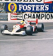 Photographie d'une Formule 1 blanche et rouge vue de trois-quarts, sur la piste de Monaco, devant des rails et des panneaux publicitaires.
