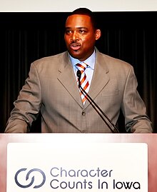 Andre Ware standing a podium in a grey suit. The podium reads "Characters Counts In Iowa".