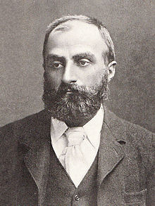 Heavily bearded man with dark receding hair, wearing a dark coloured jacket, white collar and pale tie. He is looking slightly to the left, with a solemn expression